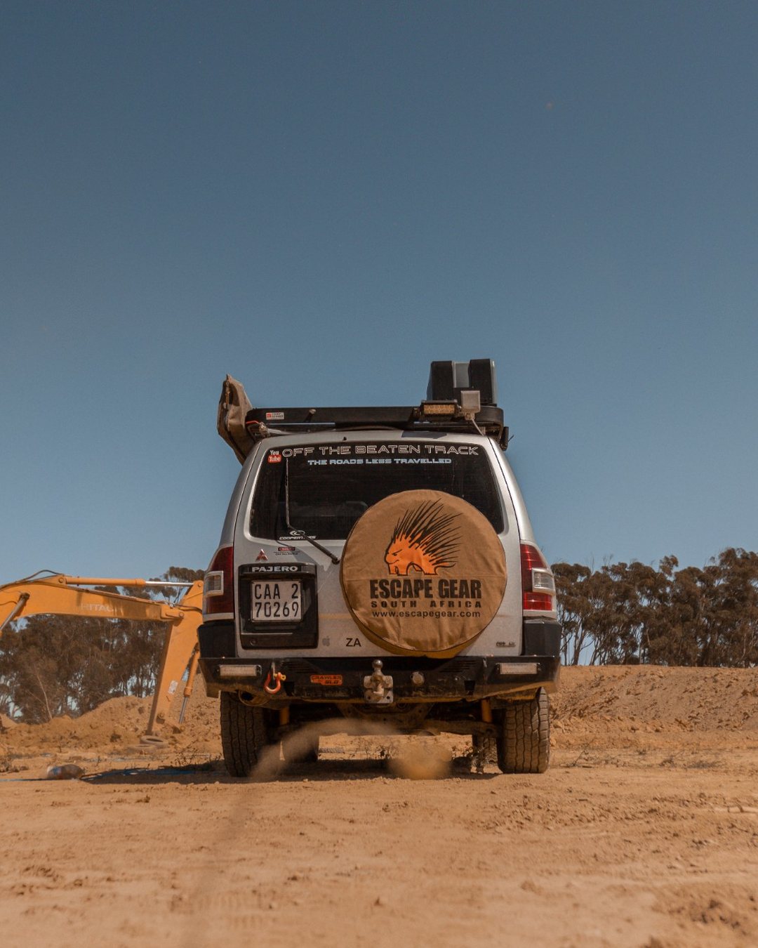 Escape Gear: a Pajero with the Escape Gear spare-wheel cover on a dirt site, an excavator behind it